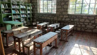 Ilustrasi: Ruang kelas kosong dengan deretan meja kayu dan rak buku di samping dinding batu.