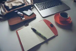 Ilustrasi: Buku catatan terbuka, pena, laptop, tas kerja, dan secangkir kopi di atas meja putih.