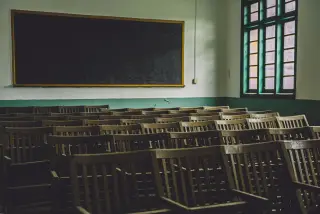 Ilustrasi: Ruang kelas kosong dengan deretan kursi kayu, papan tulis hitam, dan jendela kaca.