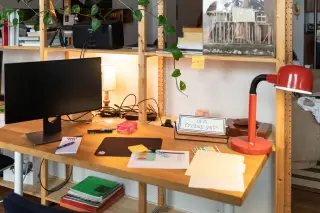 Ilustrasi: Meja kerja kayu dengan monitor, lampu meja merah, dan sejumlah berkas kertas.