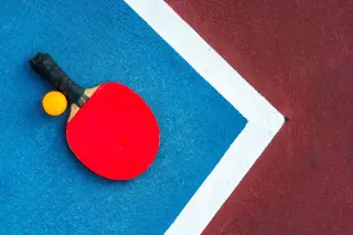 Ilustrasi: Bet tenis meja merah dan bola oranye di atas lantai lapangan berwarna biru dan merah.