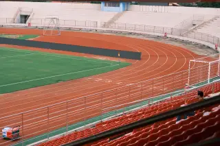 Ilustrasi: Lintasan lari atletik berwarna merah melengkung di samping lapangan rumput dan deretan kursi tribun.