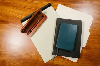 Ilustrasi: Buku catatan, kertas berkas, dan pulpen kayu di atas meja berpermukaan kayu.