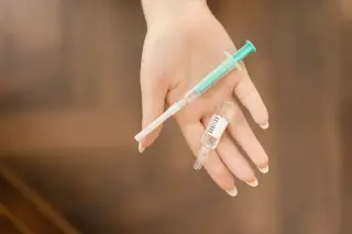 Ilustrasi: Sebuah telapak tangan memegang alat suntik medis dan sebuah ampul obat kaca.