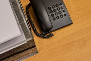 Ilustrasi: Pesawat telepon kabel hitam dan baki dokumen di atas meja kerja kayu.