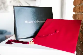 Ilustrasi: Topi toga merah dan map ijazah kelulusan diletakkan di atas meja.