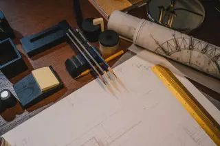 Ilustrasi: Kertas gambar teknik, pensil, dan penggaris tertata di atas meja kayu.