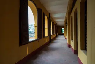 Ilustrasi: Lorong panjang berdinding kuning dengan jendela berdaun kayu yang terbuka dan lantai ubin.