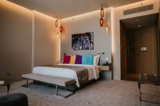 Ilustrasi: Kamar hotel modern dengan tempat tidur berhias bantal warna-warni dan lampu gantung elegan.