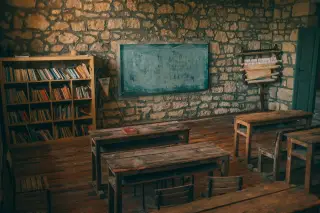 Ilustrasi: Ruang kelas kosong berdinding batu dengan deretan meja kayu dan rak buku.