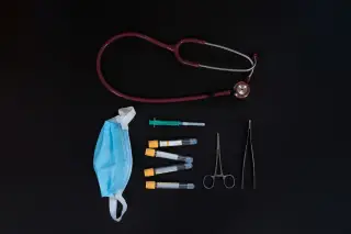 Ilustrasi: Stetoskop, masker medis, tabung sampel darah, dan alat bedah tersusun rapi di atas latar hitam.