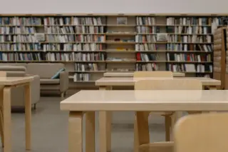 Ilustrasi: Deretan meja dan kursi kayu di dalam perpustakaan dengan latar belakang rak buku.