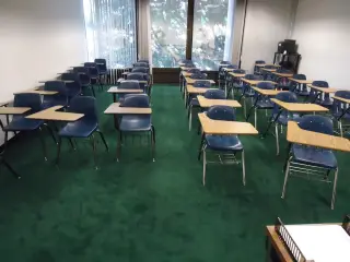 Ilustrasi: Ruang kelas kosong dengan deretan kursi belajar bertingkat dan karpet hijau.