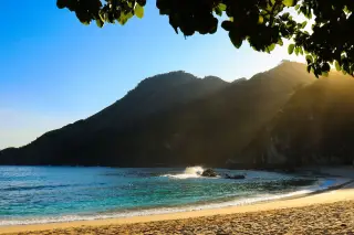 Ilustrasi: Pemandangan pantai berpasir dengan ombak tenang di dekat perbukitan hijau di bawah sinar matahari.