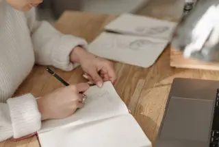 Ilustrasi: Seorang wanita menulis di buku catatan di atas meja kayu dekat laptop.