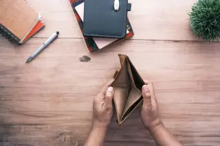 Ilustrasi: Sepasang tangan membuka dompet kulit yang kosong di atas meja kayu dengan buku catatan dan pena.