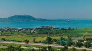 Ilustrasi: Pemandangan jalan raya di dekat pantai dengan pelabuhan dan perbukitan di kejauhan.