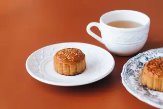 Ilustrasi: Kue bulan di atas piring keramik putih bersanding dengan cangkir teh di atas meja cokelat.