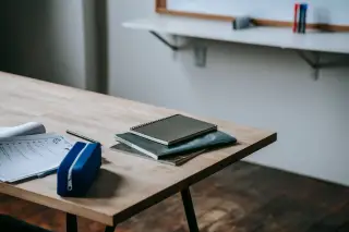 Ilustrasi: Buku catatan, lembar kerja, dan kotak pensil di atas meja kelas dekat papan tulis.