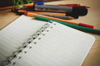 Ilustrasi: Buku catatan bergaris dengan jilid spiral terbuka di dekat beberapa pena dan pensil.