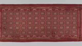 Ilustrasi: Sehelai kain tenun tradisional berwarna merah dengan pola geometris rumit terbentang horizontal.