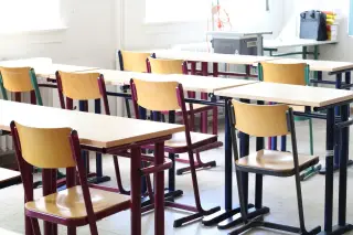 Ilustrasi: Deretan meja dan kursi kayu kosong di dalam ruang kelas berpenerangan alami.
