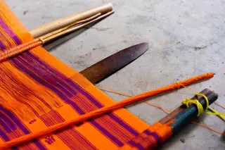 Ilustrasi: Kain tenun warna oranye dan ungu terpasang pada alat tenun tradisional di lantai semen.
