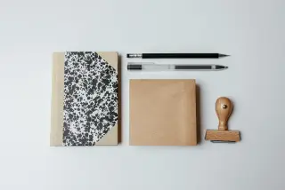Ilustrasi: Buku catatan, alat tulis, kertas cokelat, dan stempel kayu di atas latar putih.
