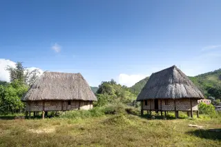 Ilustrasi: Dua rumah panggung tradisional beratap jerami dan berdinding anyaman di area perbukitan.
