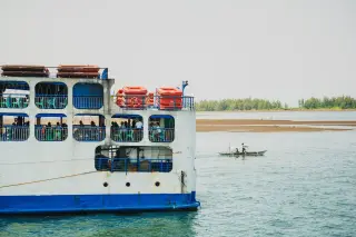 Ilustrasi: Sebagian badan kapal feri penumpang berlayar di laut dekat perahu kecil dan garis pantai.