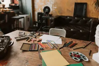 Ilustrasi: Pensil warna, kertas gambar, dan palet cat di atas meja kayu dalam sebuah ruangan seni.