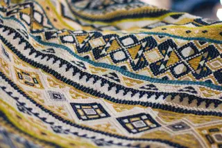 Ilustrasi: Kain tenun dengan pola geometris berwarna kuning, biru tua, dan putih.