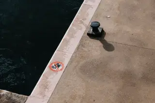 Ilustrasi: Bollard tambatan kapal dan simbol larangan di tepi dermaga beton dekat air laut.