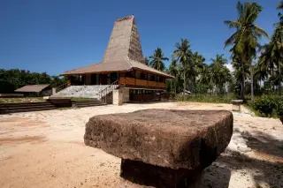 Ilustrasi: Batu kubur megalitik di latar depan dengan rumah adat beratap menara khas Sumba di bawah langit biru.