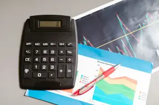 Ilustrasi: Kalkulator hitam, pulpen, dan cetakan grafik keuangan di atas meja.