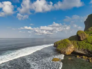 Ilustrasi: Pemandangan tebing karang hijau di tepi laut berombak di bawah langit biru berawan.