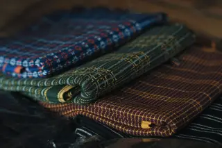 Ilustrasi: Tumpukan kain tradisional bermotif dengan berbagai warna tersusun rapi.