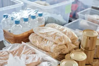 Ilustrasi: Bungkusan pasta kering, botol air mineral, dan kaleng makanan tersusun rapi di wadah plastik.