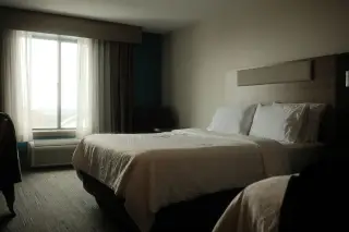 Ilustrasi: Kamar hotel dengan tempat tidur tertata rapi di samping jendela berhordeng tipis.