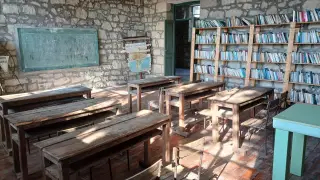 Ilustrasi: Ruang kelas berdinding batu dengan deretan meja kayu kosong dan rak buku perpustakaan.