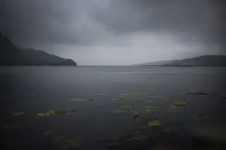 Ilustrasi: Pemandangan perairan tenang berlatar perbukitan di bawah langit mendung gelap kelabu.
