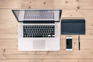 Ilustrasi: Laptop, buku catatan, pena, dan ponsel pintar tertata rapi di atas meja kayu.
