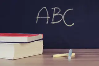 Ilustrasi: Buku bertumpuk dan kapur tulis di atas meja dengan latar papan tulis bertuliskan ABC.