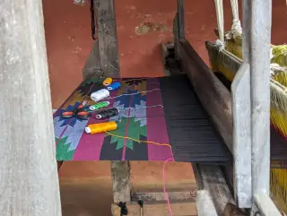 Ilustrasi: Alat tenun kayu tradisional dengan kain bermotif warna-warni dan beberapa gulungan benang.