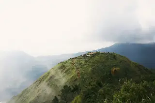 Ilustrasi: Pemandangan bukit hijau berawan dengan jalur setapak tanah menuju puncak yang ada tenda kemah.