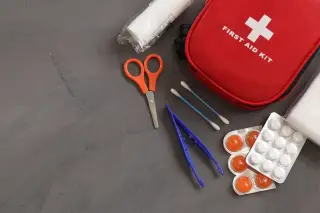 Ilustrasi: Kotak pertolongan pertama merah dengan gunting, pinset, obat, dan perban di atas meja.