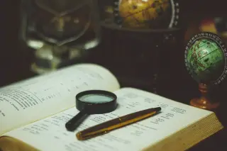 Ilustrasi: Buku terbuka dengan kaca pembesar dan pena di atasnya serta miniatur bola dunia.
