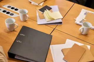 Ilustrasi: Meja rapat kayu dengan map binder hitam, tumpukan berkas dokumen, buku catatan, dan cangkir putih.