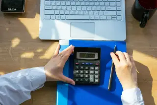 Ilustrasi: Sepasang tangan di meja kerja memegang kalkulator di dekat laptop dan buku catatan.
