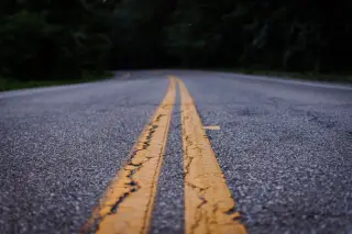 Ilustrasi: Jalan aspal lengang dengan marka kuning ganda di tengah melintasi area berpohon.